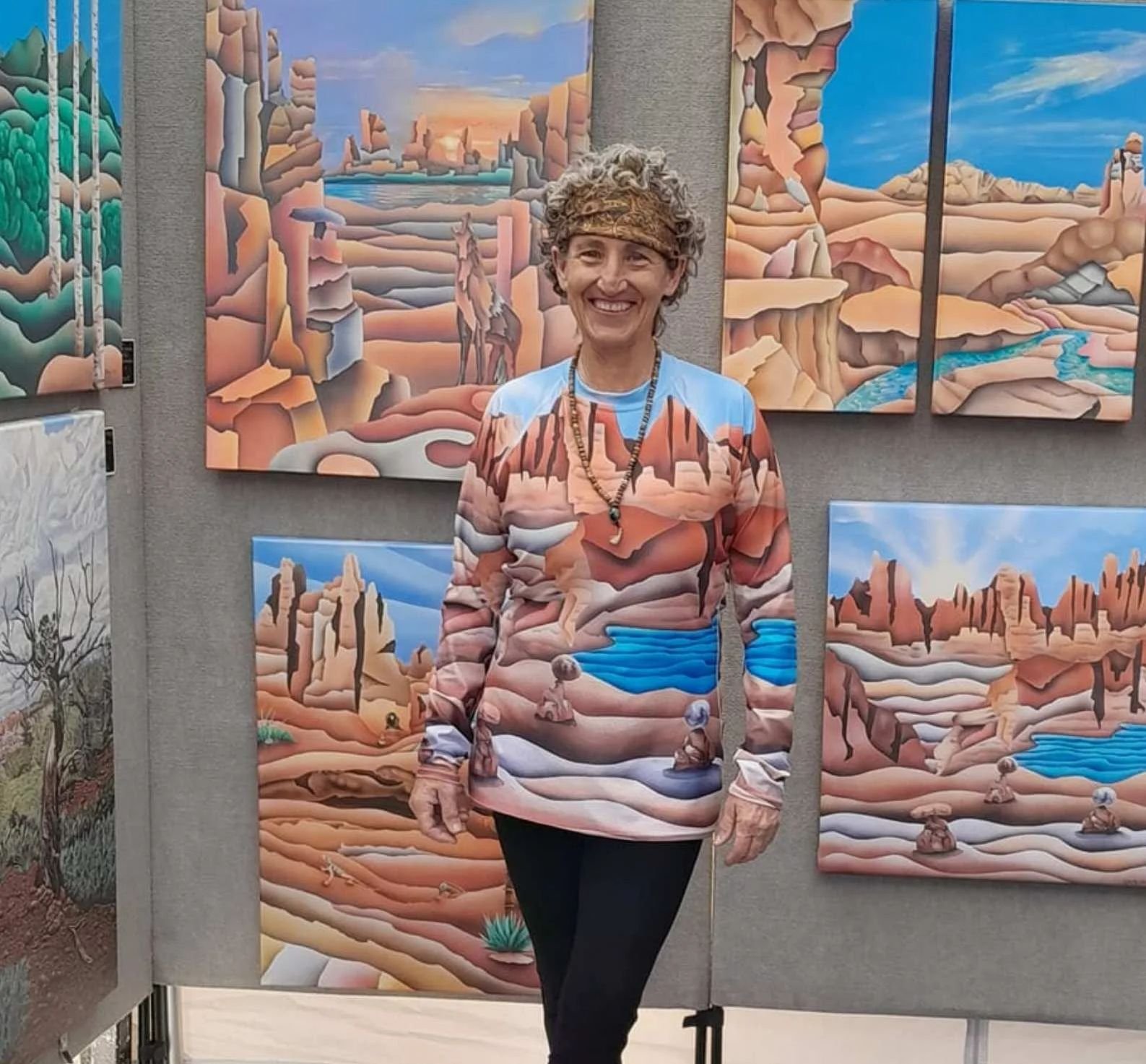 Artist Therese Tice standing in front of her desert landscape paintings while wearing a matching hand-painted desert-themed shirt at her art booth.